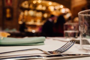 Glasses, fork, knife served for dinner in restaurant with cozy interior.Blurred people enjoying food
