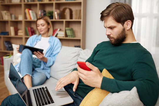 Couple Using Gadgets At Home. Married Couple Of Freelancers Work From Home. Guy And Girl In Casual Clothes With Gadgets Work In The Living Room At The Sofa 