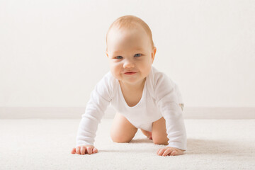 Happy smiling adorable baby boy in white bodysuit crawling on knee and arms on light beige home...
