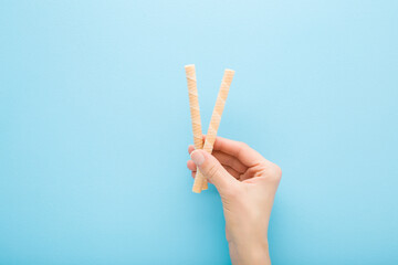 Young adult woman hand holding two striped white vanilla wafer sticks on light blue table background. Pastel color. Closeup. Sweet snack. Top down view.