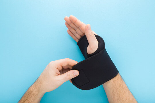Young adult man hand putting protective black dark elastic wrist bandage on light blue table background. Pastel color. Top down view. Closeup. Point of view shot.