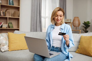 Positive Woman Holding Credit Card. Smiling Woman Paying Online, Using Laptop, Holding Plastic Credit Card, Sitting On Couch At Home. Young Female Shopping, Making Secure Internet Payment 