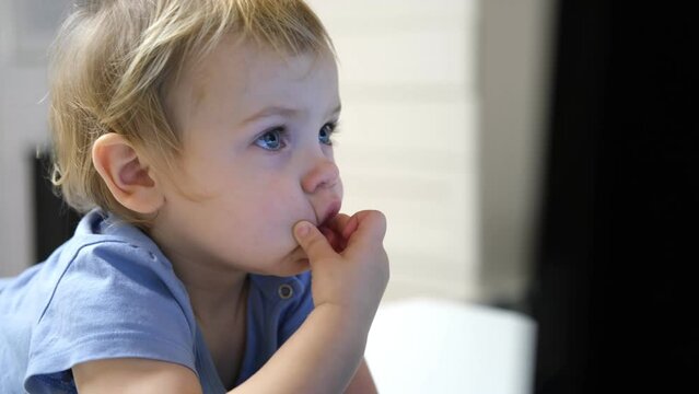 Little girl Stares Intently at the Monitor. Small One Years Old Child Watching Laptop Screen and Making Faces. Toddler Sitting at Table and Looking into Computer on Children's Cartoons, Video or TV.v