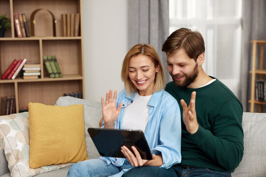 Couple Talking Tablet At Home. Young Couple Using Digital Tablet While Sitting On Couch. Happy Smiling People Watching Video On Digital Tablet At Home 