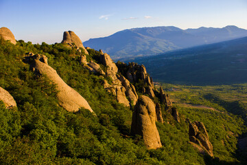 Fototapeta na wymiar The slope of Demerdzhi Mountain with stones and large boulders covered with trees, bushes and greenery with distant hills in a haze and villages in a valley in the summer in Crimea in Ukraine 