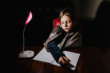 Blackout in Ukraine. A Ukrainian, a little girl, a child, a schoolgirl sits at a table in the dark without light with a lighting lamp and does her homework.