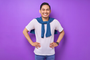 Smiling young Asian man wearing white t-shirt posing holding hands on his waist and confident at the camera isolated over purple background. people lifestyle concept