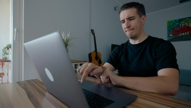 Focused programmer develops new video game on modern laptop. Man types on gadget sitting at wooden table in apartment living room slow motion