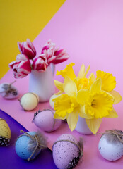 Two vases with flowers next to decorated Easter eggs on a multi-colored background copy space