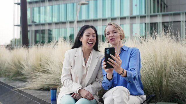 Cheerful women coworkers sharing video call with professional colleague friend using smartphone sitting on office garden