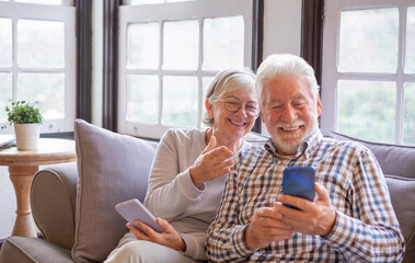 Portrait of cheerful senior couple while sitting together on sofa laughing. Elderly happy couple relaxing while using smartphones