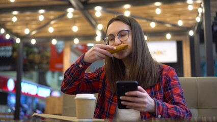 A young woman eats pizza in a cafe with a phone in her hand. A girl in a red shirt and glasses is watching memes on a smartphone.