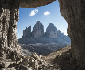 Drei Zinnen in den Dolomiten durch ein Felsfenster mit Wolken und blauem Himmel.