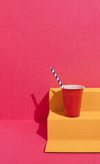 Yellow paper podium with red plastic cup and paper straw on magenta background. Minimal concept.