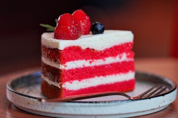 A slice of red velvet cake with cream cheese frosting, garnished with strawberries and blueberries, served on a plate with a fork.