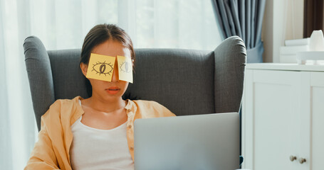 Young asia female girl or university student sit on sofa chair with laptop feel sleepy, tired put sticky note fake eyes take a rest from overwork in living room at home. Work life balance concept.