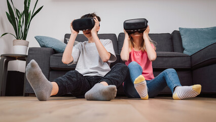 Fototapeta premium Two children, a boy and a little girl wearing VR headsets sit in the living room playing, having fun in a virtual world through an interactive experience