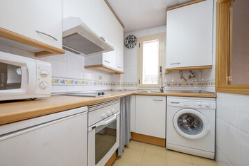 Corner of a kitchen furnished with white furniture and a wooden countertop with integrated white appliances and a border on the tiles