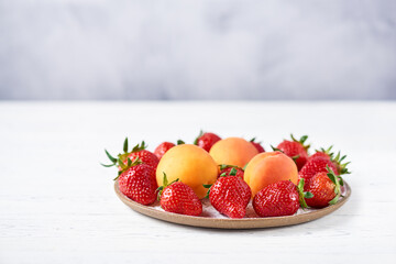 Ripe strawberries and apricots on a flat ceramic plate on a white wooden table. Copy space, close-up.