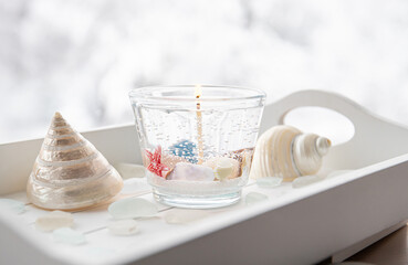 Homemade underwater seashell clear gel wax candle inside clear cup. Beautiful candle on white wood tray with sea glass and seashells on window sill at home.