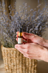 Woman's hands are holding a bottle with a pipette with lavender oil into the bottle on lavender background in straw basket