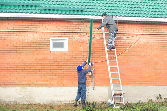 Two Men Prepare Their House For A Snowy Winter, They Install Snow Traps On The Roof With Their Own Hands From A High Ladder