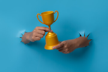 Female hand holding a champion golden trophy breaks through blue paper background.