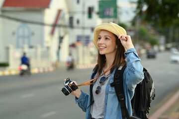 Fototapeta na wymiar Young female tourist with backpack and digital camera standing on city street. Blogger, vacation and journey concept