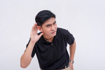 An annoyed and agitated young man leans forward to hear someone better. Having difficulty hearing, partially deaf, or trying to listen to inaudible speech. Isolated on a white background. - obrazy, fototapety, plakaty