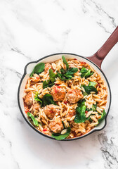 One pot lunch. Orzo pasta with meatballs and spinach in tomato sauce in a frying pan on a light background, top view