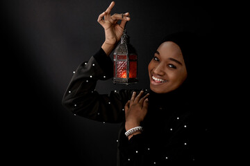 Happy african muslim woman in black hijab holding celebratory lamp and smiling at camera, Ramadan lantern on black background
