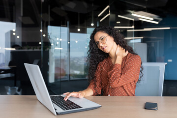 Fototapeta na wymiar Overtired business woman working inside office with laptop, Hispanic female worker has severe neck pain, massaging muscles with hand.