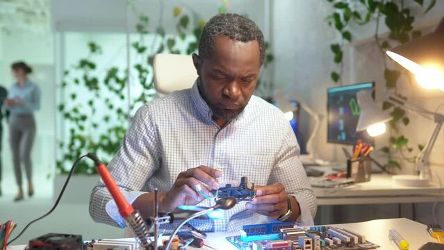 Portrait of focused professional young man working in service setting up cooler on motherboard. Concentrated African engineer repairing computer sitting in modern office using tools.