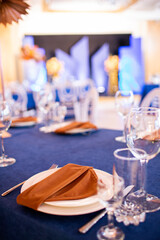 Festive table with blue cloth, napkins, plates, cutlery and glasses prepared for wedding guests in restaurant. Beautiful floral arrangement in gold metal vase with number three. Amazing servings