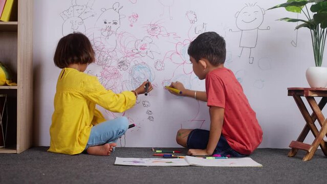 Young siblings kids drawing with crayon color on wall by looking at camera at home - concept of mischief, troublesome children and childhood lifestyles.