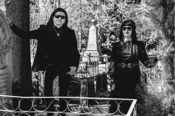 Stylish dark gothic couple with black clothes between graves in cemetery in a sunny day (in black...