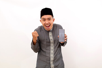 Happy asian muslim man standing while holding a cellular phone and clenching hand. Isolated on white background