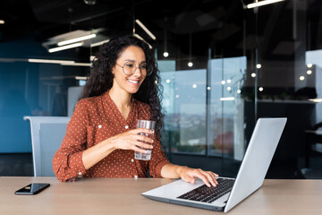 Fototapeta na wymiar Successful and happy Latin American business woman at work inside the office, female worker holds a glass of clean water in her hands and drinks, businesswoman is satisfied achievement, uses a laptop.