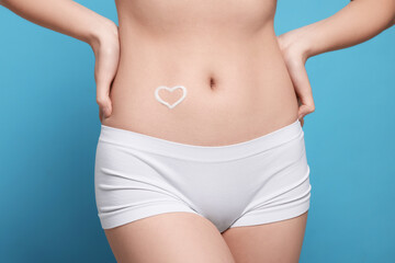 Woman with heart made of body cream on her belly against light blue background, closeup
