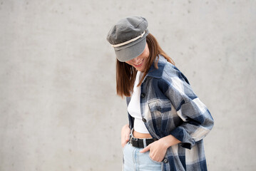 Girl in black and white checkered shirt, sailor's hat, cement wall in the background. Gesture that...