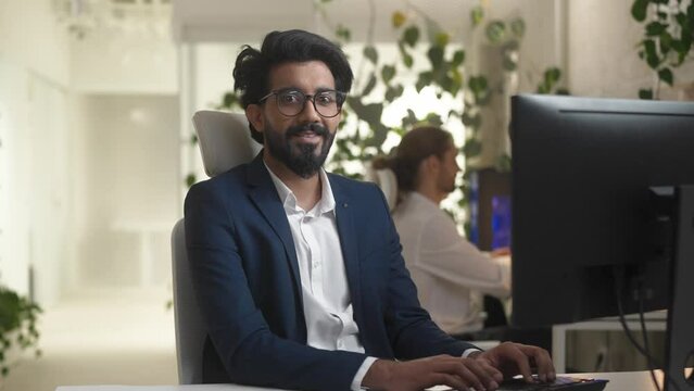 Portrait of young professional Indian IT company worker typing on keyboard using computer in modern office. Focused bearded happy man developer coding wearing suit looking at camera smiling.