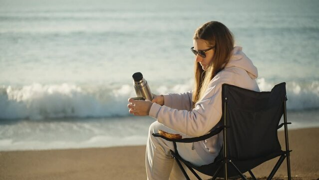 A girl on a Chair by the sea Pours hot tea from a Thermos. Sunrise early in the morning.
