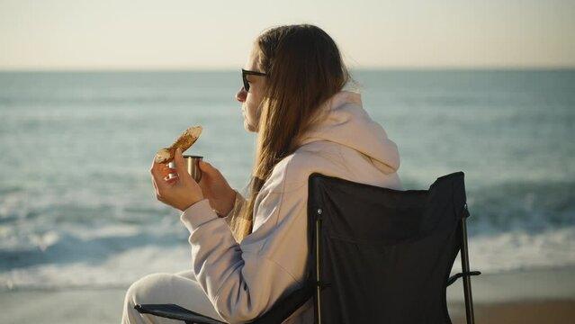 It's a cool morning, a girl is sitting on a chair on the sea Beach, eating a bagel and writing down Hot Tea. Admiring the sunrise.