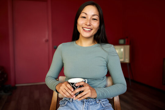 A relaxed woman is sitting with a glass of coffee or tea or a hot drink, looking at the camera, wearing a blue sweater with long arms, smiling gently. Half-length portrait of a girl at a job interview