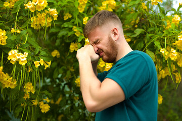 Portrait of handsome young allergic man is suffering from pollen allergy or cold on natural flowers, flowering tree background at spring or sunny summer day, sneezes, blowing his runny nose rubs eyes