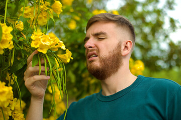 Portrait of handsome young allergic man is suffering from pollen allergy or cold on natural flowers, flowering tree background at spring or sunny summer day, sneezes, blowing his runny nose rubs eyes