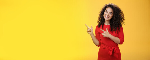 Friendly cute assertive female assistant in elegant red dress with curly hair tilting head flirty and smiling broadly, satisfied as pointing at upper left corner, recommending product over yellow wall