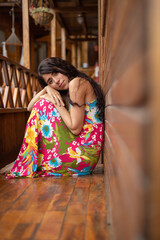 Fototapeta premium posing sitting inside a wooden house a young latin woman wearing a colorful dress and with long hair, lifestyle and natural beauty in the daytime, model portrait