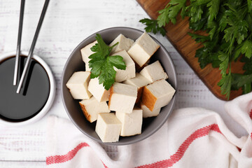 Bowl of smoked tofu cubes, soy sauce and parsley on white wooden table, flat lay
