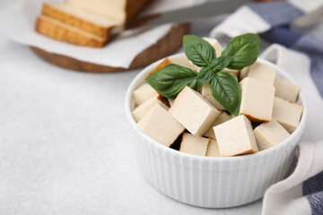 Bowl of smoked tofu cubes with basil on white table, closeup. Space for text
